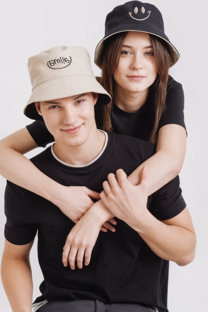 Two people wearing black t-shirts and bucket hats with a smiley face design on a white background.