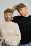 A man and a woman wearing matching olive green knit beanie hats, posed against a light gray studio background. The woman wears a cream knit sweater and blue jeans, while the man wears a black sweatshirt and blue jeans.