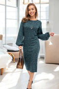 A woman wearing a dark teal heathered knit long-sleeve midi dress, holding a brown patterned handbag in a bright, modern living room.