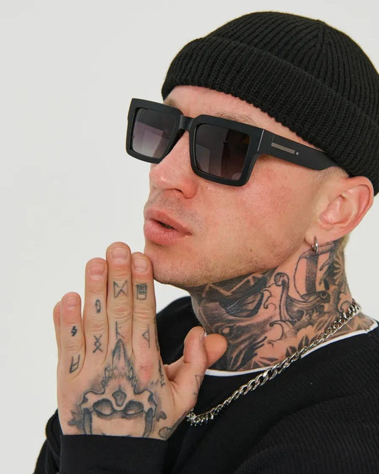 Person wearing sunglasses, black beanie, and black clothing with visible tattoos on a plain background