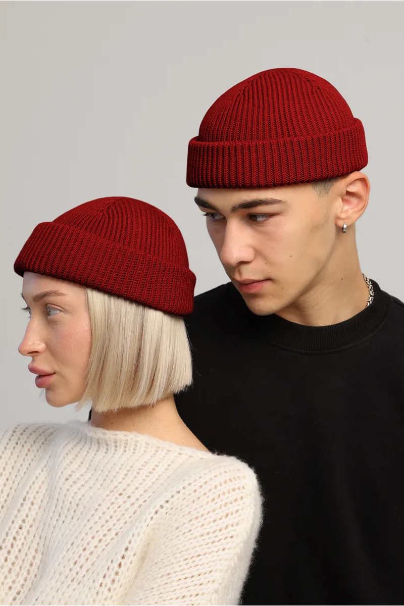 Two models wearing matching deep red ribbed knit beanie hats, posed against a light gray studio background.
