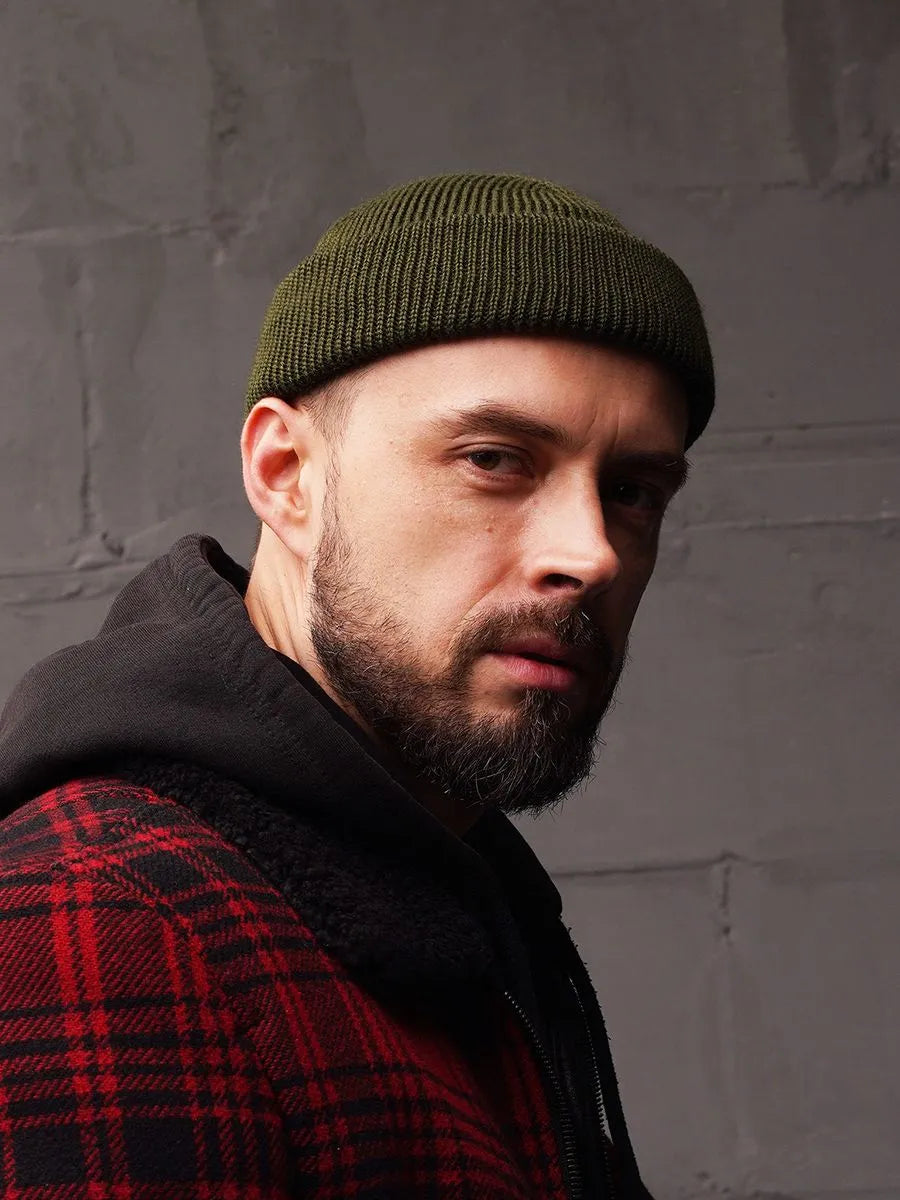 Man wearing a green beanie and red plaid coat against a gray brick wall