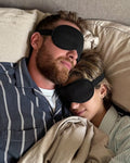 Two people lying in bed under a blanket, wearing eye masks.