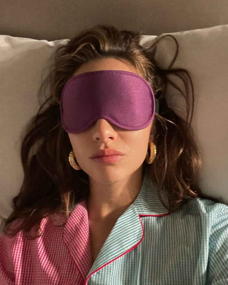 Person wearing a purple sleep mask and pajamas lying on a bed.