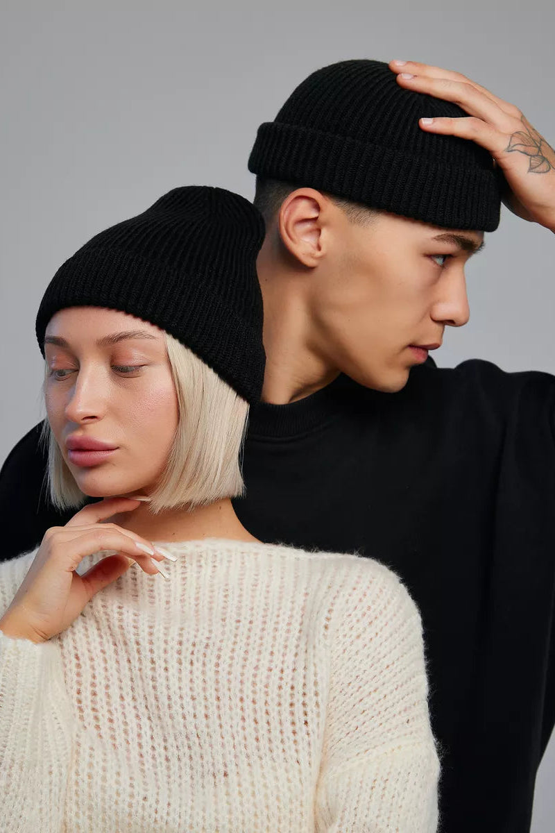 Two models wearing black ribbed knit beanie hats, one in the foreground with short platinum blonde hair wearing a cream knit sweater, and one behind with dark hair wearing a black long-sleeve top.