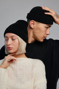 Two models wearing black ribbed knit beanie hats, one in the foreground with short platinum blonde hair wearing a cream knit sweater, and one behind with dark hair wearing a black long-sleeve top.