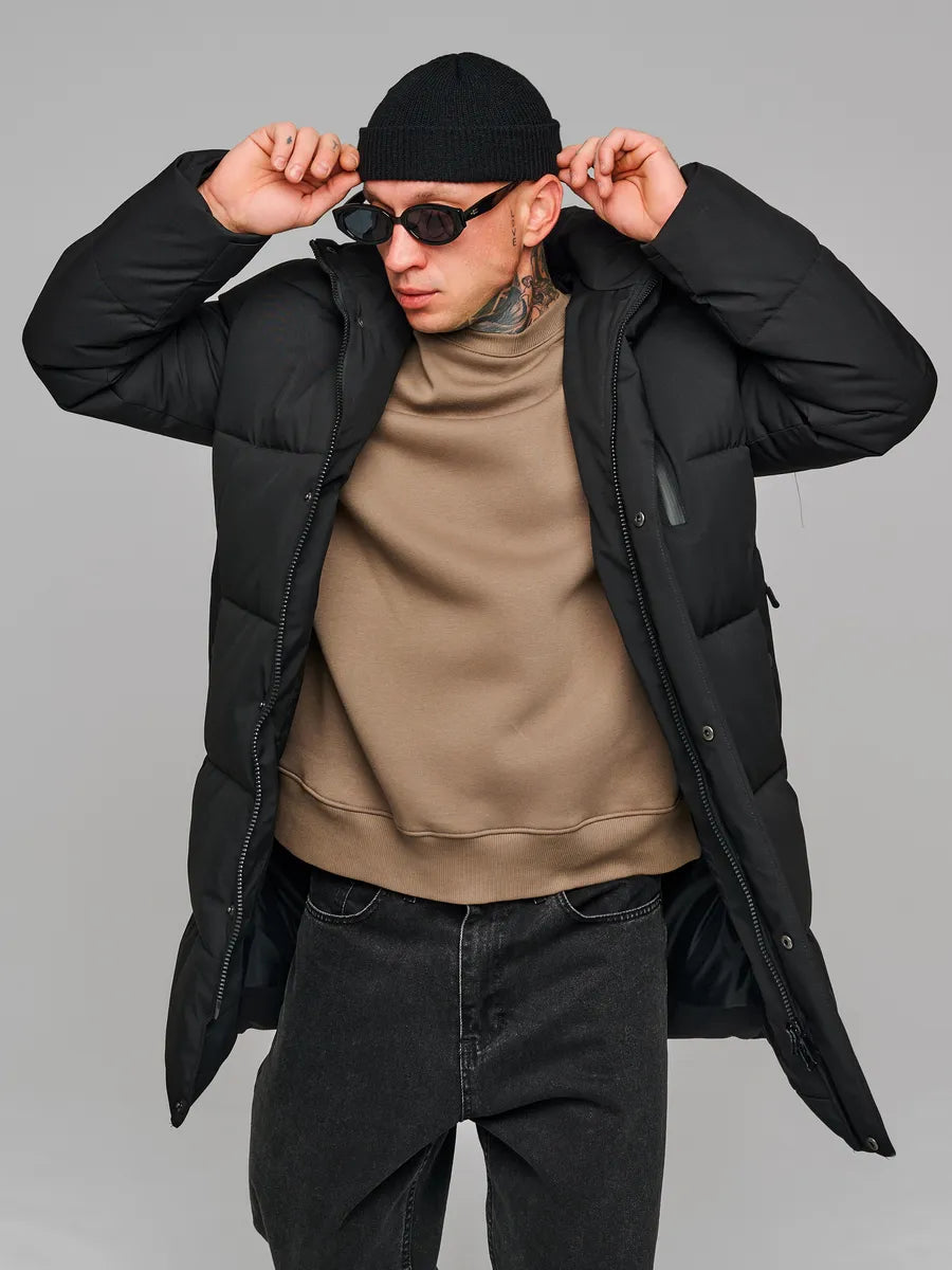 Man wearing a black puffer jacket, beige sweater, and sunglasses on a gray background