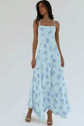 A woman modeling a light blue maxi dress with a blue floral print, featuring thin spaghetti straps and a flowing skirt, set against a plain white studio background.
