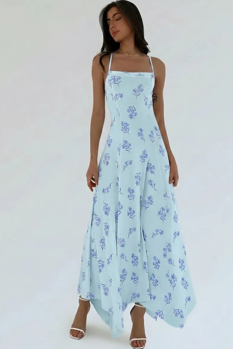 A woman modeling a light blue maxi dress with a blue floral print, featuring thin spaghetti straps and a flowing skirt, set against a plain white studio background.