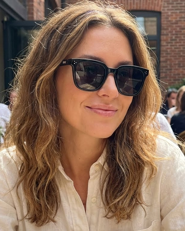 Woman wearing sunglasses in an outdoor setting with people and plants.