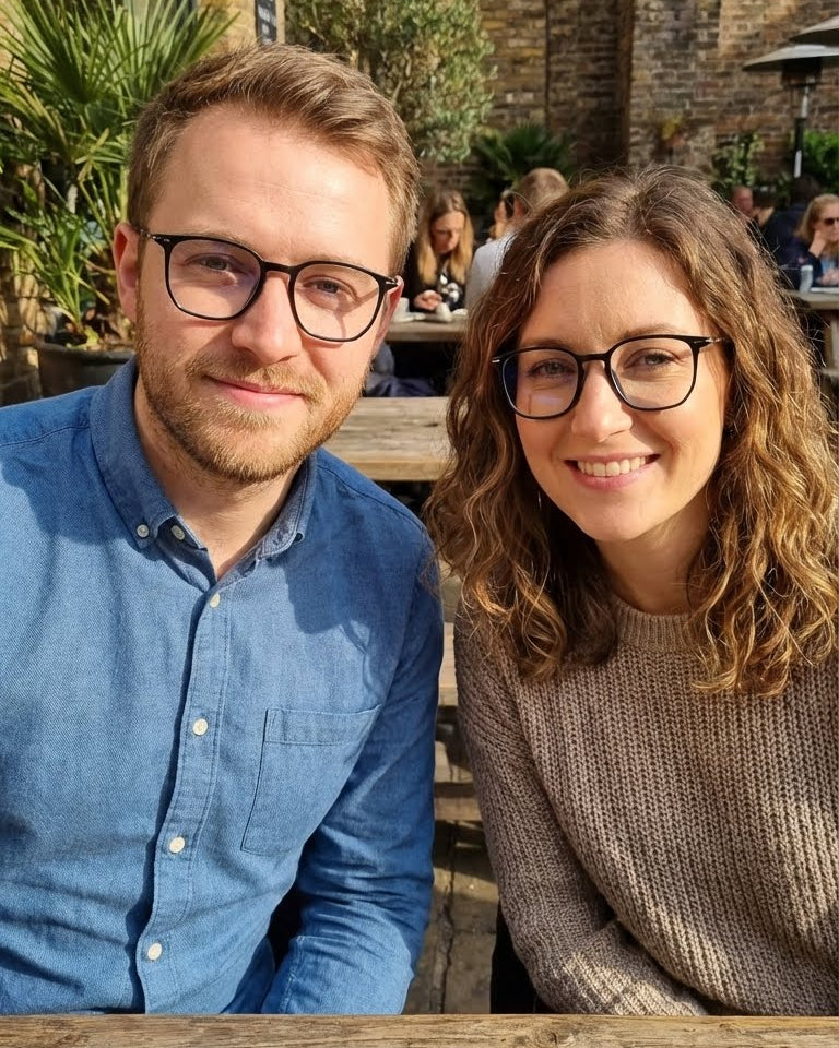 Two people sitting at an outdoor table with a blurred background of people and buildings.
