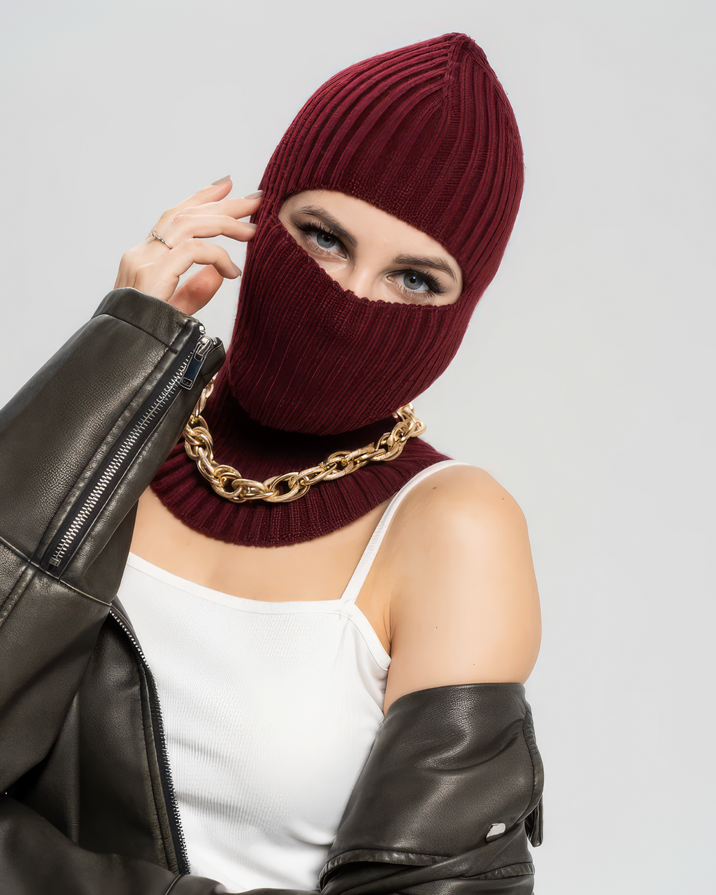 Person wearing a burgundy knitted balaclava with a gold chain, leather jacket, and white top on a light gray background.
