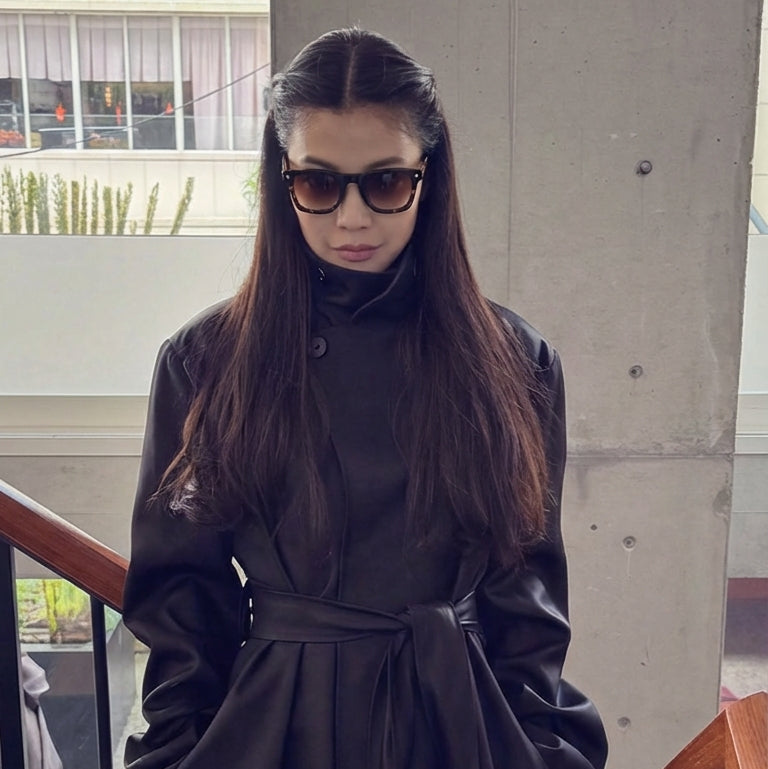 Person wearing a long black coat and sunglasses indoors.