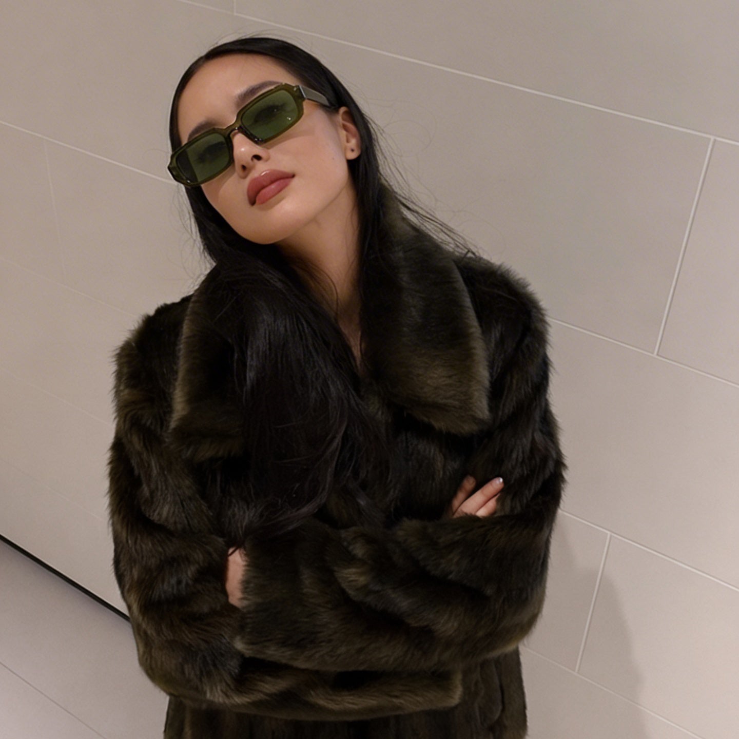 Woman wearing a dark fur coat and sunglasses against a light-colored wall.