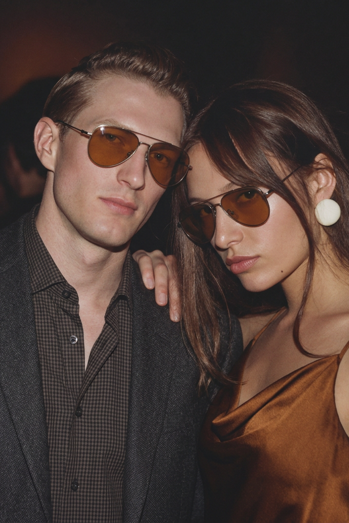 Two people wearing sunglasses posing together in a dimly lit setting.